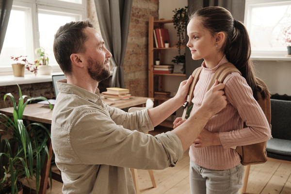 Dad with a beard in a light shirt over a T-shirt helps his daughter with long tidied up hair to put a school bag on her shoulders