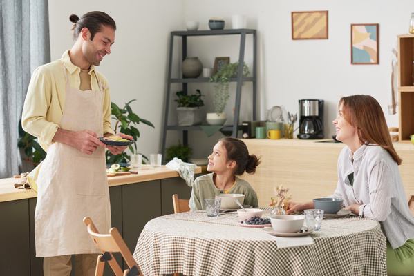 A smiling man in a yellow shirt and apron serves breakfast to the table where his smiling wife and daughter sit