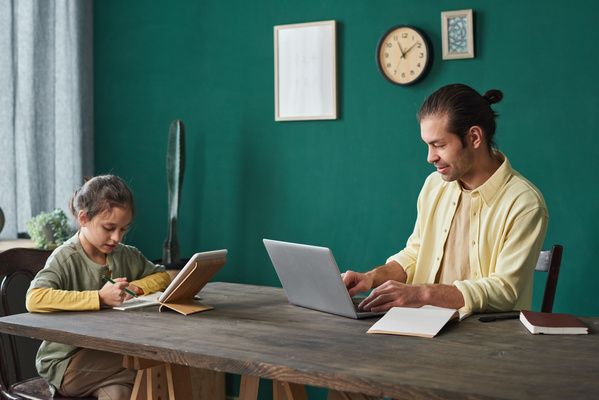 A busy father with his tidied up hair dressed in a shirt working on a laptop at a table and his daughter doing homework using a tablet