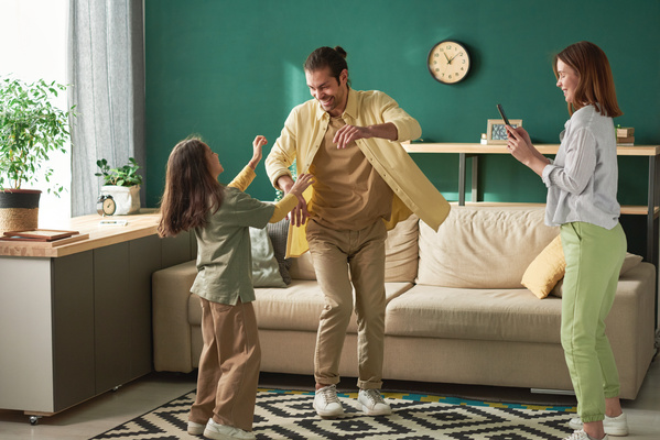 A woman filming on the phone her husband with tidied up hair dressed in a yellow shirt dancing with their daughter