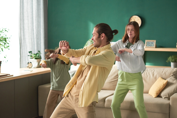 A man in a yellow shirt dances with his wife in green pants and daughter in a bright room with an emerald-colored wall