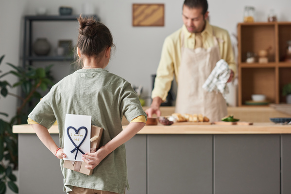 A girl with her hair in a bun holding a homemade Fathers Day card and a gift behind her back goes to congratulate her father cooking in the kitchen