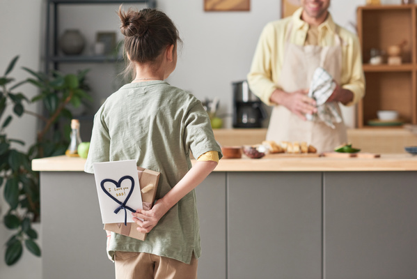 A girl holding a gift and a homemade postcard for Fathers Day being ready to congratulate her dad cooking in the kitchen