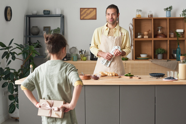 A father in an apron cooking in the kitchen looks at his daughter holding a handmade Fathers Day card behind her back for him
