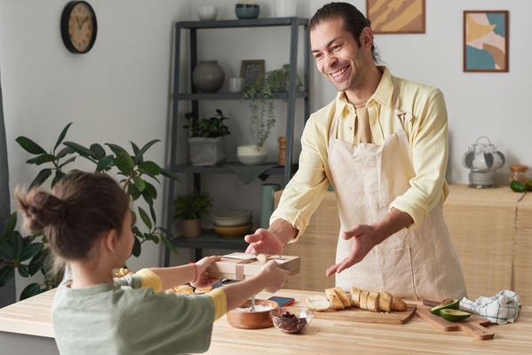 A father in an apron and with a kitchen towel in his hands smiling at his daughter holding a gift behind her back to congratulate him in the kitchen