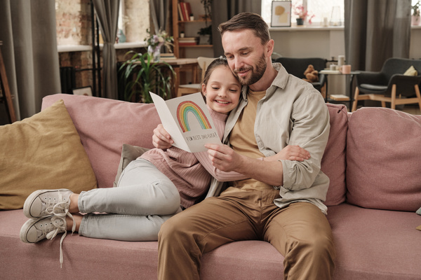 A father sitting in his daughters arms on a pink sofa reads congratulations on a homemade Fathers Day postcard from her