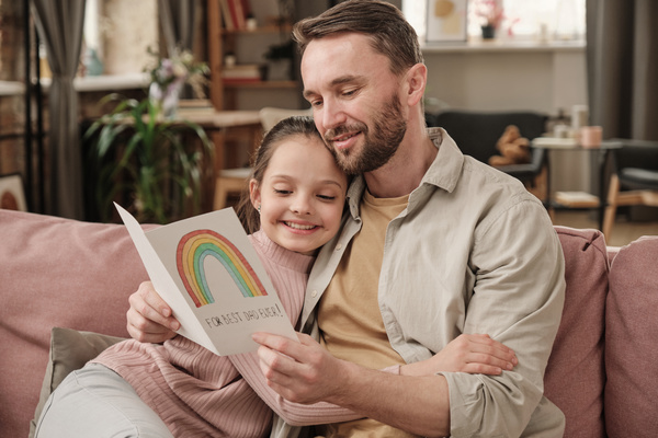 A man sitting on a pink couch reading congratulations on a greeting Father's Day card and hugging his daughter