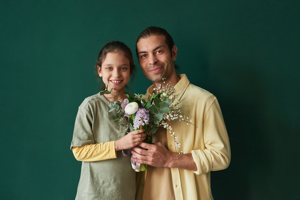 A man in a yellow shirt and a smiling daughter with tidied up hair hold a bouquet of flowers in their hands standing against a dark green background