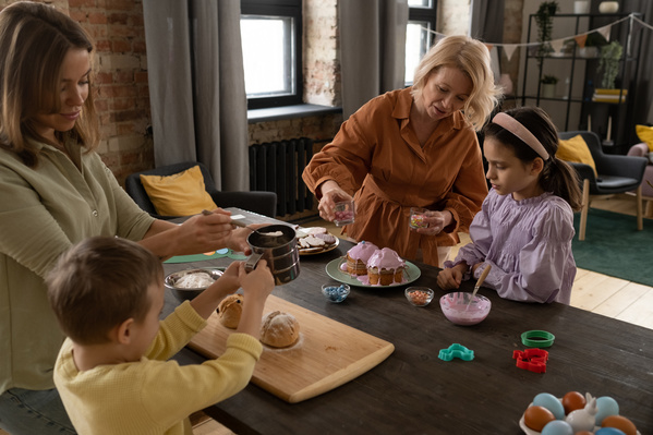 A family consisting of two children a mother and grandma decorating Easter cakes in pink icing and buns with powdered sugar with sweets standing around a wooden table in the kitchen
