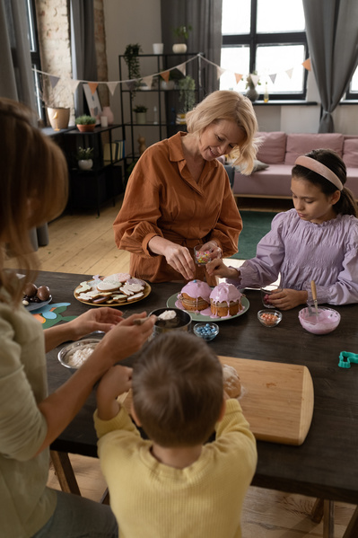 A grandma with blonde hair and a granddaughter in a pink blouse decorating an Easter cake in pink icing with sweets standing at the table with mom and her son