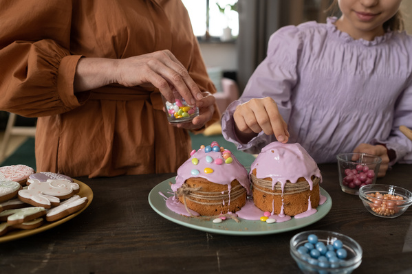 Easter pastry in pink glaze is decorated with colorful sweets by grandma and granddaughter standing at a dark wood table