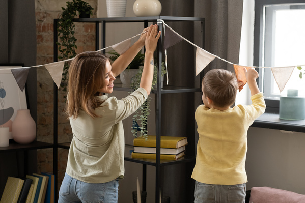 A woman in a light shirt smiling hangs holiday flags garland in the room with her son in a yellow sweater