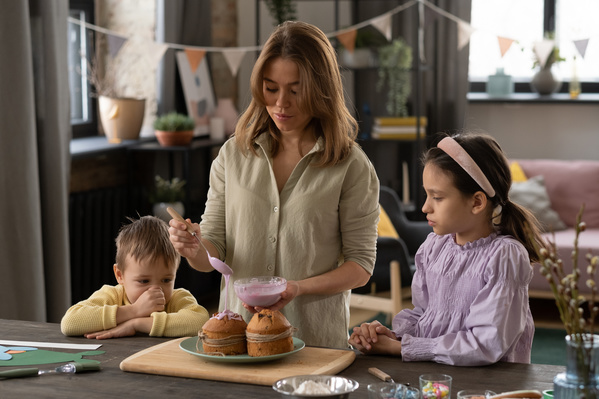 A woman dressed in a light shirt surrounded by her children watching with interest as she decorates with pink icing from a glass cup an Easter cake on a green plate