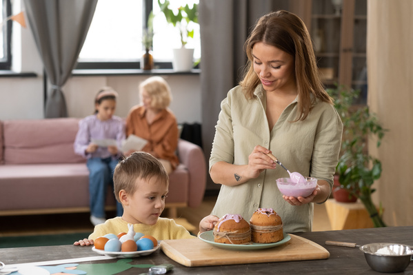 A woman dressed in a light shirt and her son are about to glaze the top of the Easter cake on the kitchen wooden table with pink icing