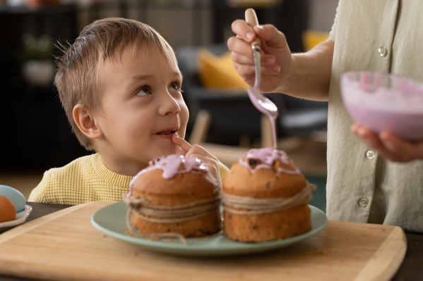 A boy in a yellow sweater watches with interest his mother glazing an Easter cake with pink icing