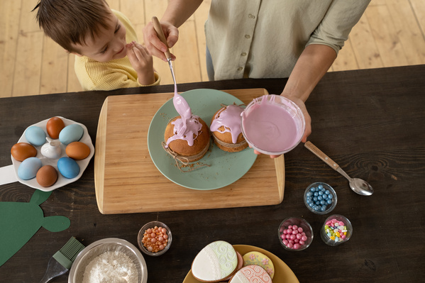 Top view of a boy in a yellow sweater watching his mother in a pistachio-colored shirt smearing Easter cakes with pink icing