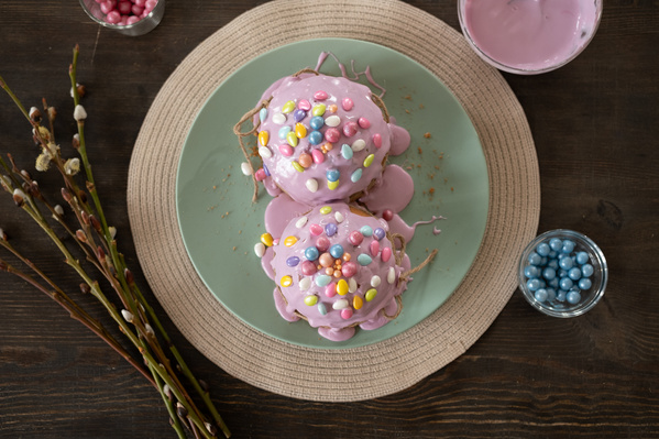 Top view of Easter pastries decorated with pink icing and colored sweets on a pistachio-colored plate standing on a table with willow twigs