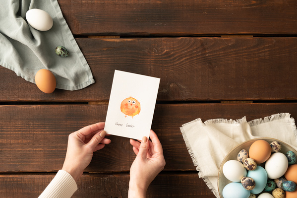 Top view of chicken and quail Easter eggs on white and blue linen napkins and a themed postcard with a drawing in hands on a dark wood table background