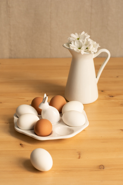 A white plate of hen eggs decorated with a rabbit figure and a jug with camomiles on a wooden table
