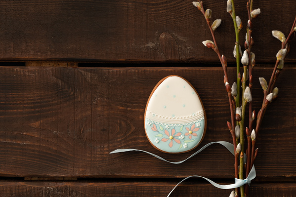 Easter egg-shaped treat in white and blue coloring with a thematic pattern and willow twigs tied with a blue ribbon on a dark wood surface