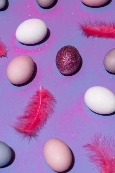 Colored Easter chicken eggs are spread on a purple surface with sparkles and pink feathers