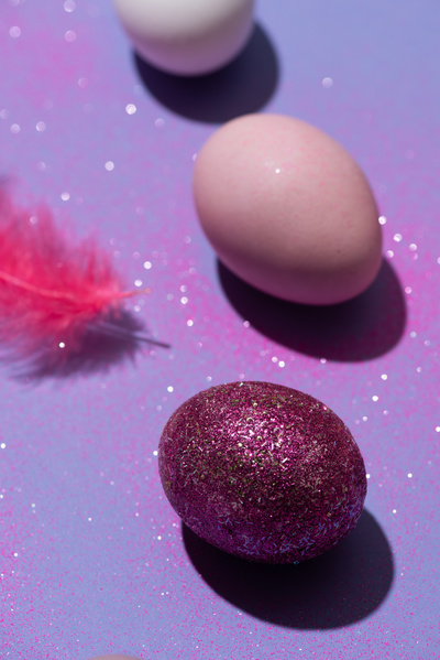 Easter egg covered with pink sparkles on a purple surface with a pink feather and eggs