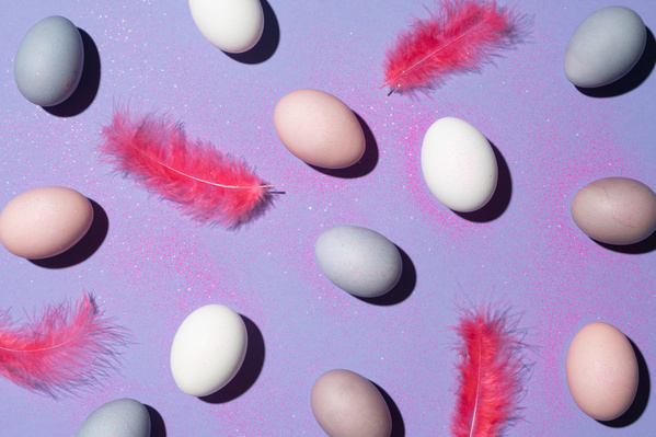 A flatlay of Easter eggs colored in different colors with pink feathers on a purple background in pink sparkles
