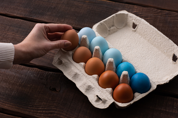 An egg is taken from carton with blue and brown Easter eggs on a dark wood surface