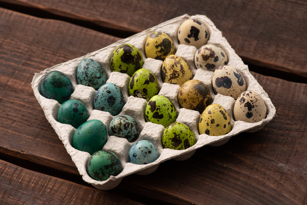 Carton on a dark wooden table filled with quail Easter eggs painted in different colors