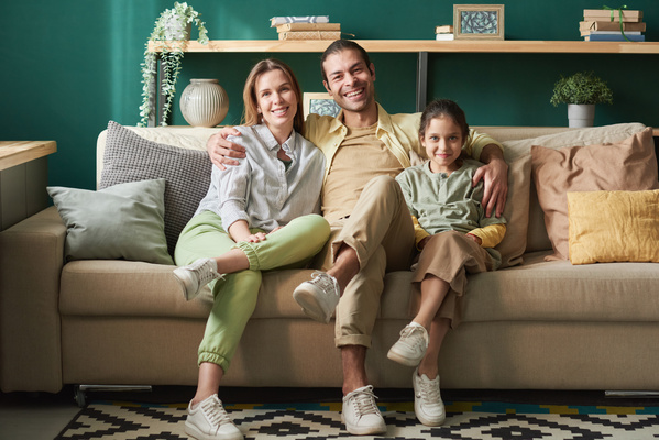 A man in a yellow shirt sitting on the sofa and hugging his wife and daughter who have crossed their legs in a bright living room
