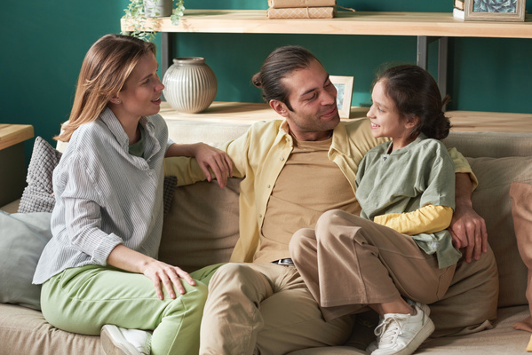 A family photo of a man in a yellow shirt sitting in the living room on a carpet with a geometric pattern surrounded by a beautiful wife and a smiling daughter with her hair down