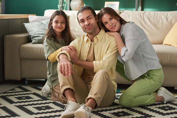 A family photo of a man in a yellow shirt sitting in a bright room on a carpet with a geometric pattern with his wife resting her head on his shoulder and a smiling daughter with her hair down
