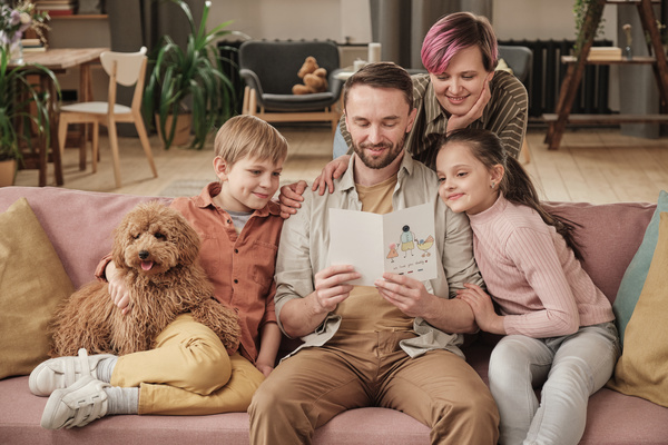 A man sitting on a sofa with his son holding a dog and his daughter while their mother standing behind and reading a greeting card in a homemade postcard with an illustration for Fathers Day