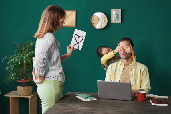 A woman is going to congratulate her husband on Fathers Day and present a postcard with an application to him sitting at a table with a laptop while his daughter covers his eyes with her hands