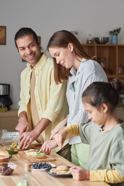 A man slices avocado cooking breakfast together with his wife and daughter standing at the kitchen table