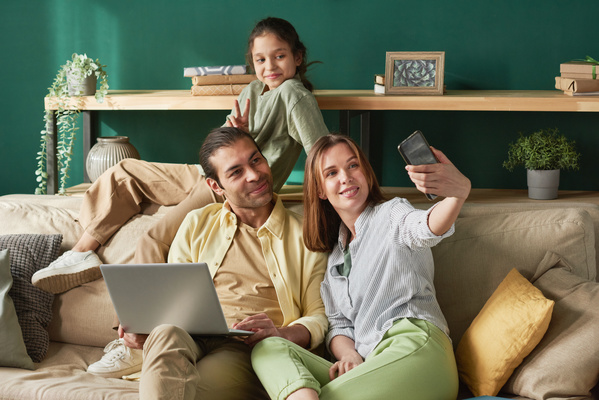 A woman sitting on the sofa in the living room with her husband holding a laptop and posing daughter takes a selfie