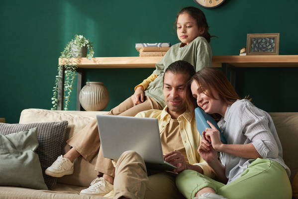 A family consisting of a father in a yellow shirt a mother holding a blue book and a daughter with her hair in a bun spending time on the sofa in the living room with a laptop