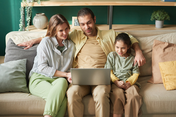 A man sitting on the sofa in the living room with his daughter in a green long-sleeved T-shirt and his wife in a striped shirt watching a movie on a silver-colored laptop