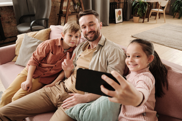 A girl with her hair in a ponytail taking a photo on the phone with her dad and brother in a red shirt while sitting on a pink sofa