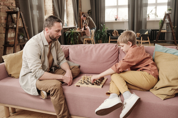 A father in a light shirt sitting on a pink sofa playing chess with his blond son
