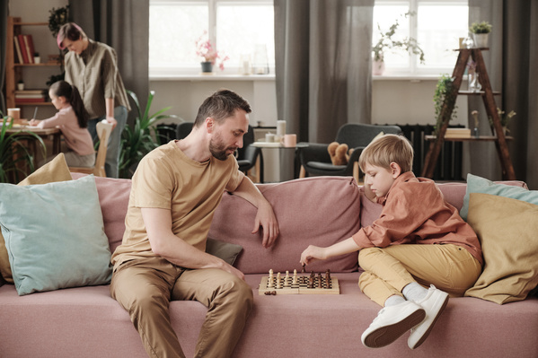 A man in a light shirt plays chess with his blond son on a pink couch