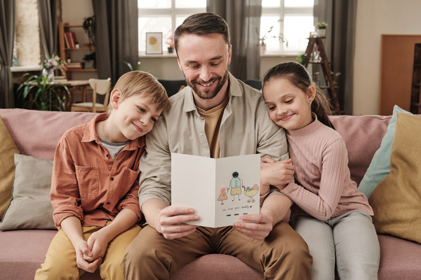 A man holding a Fathers Day card from his son with blond hair and his daughter with a ponytail sitting on the couch together