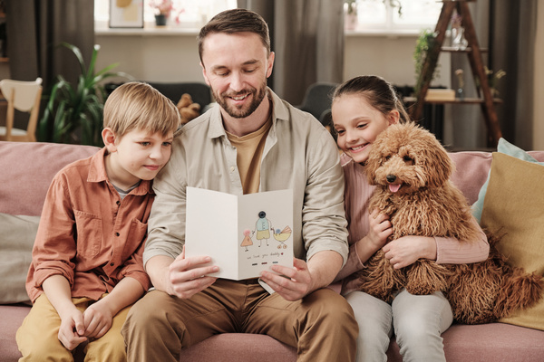 A father with a homemade postcard in his hands given to him by his children sitting with him and their dog on the couch