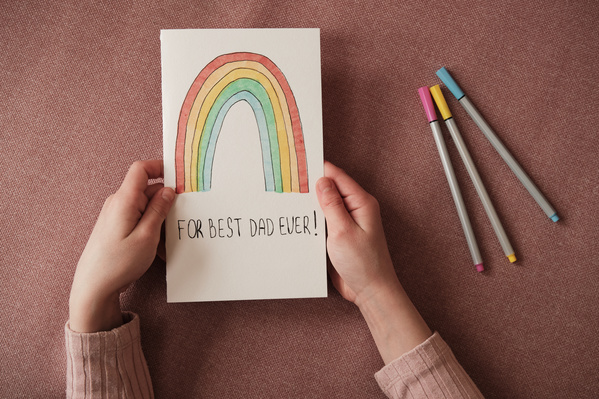 A homemade Fathers Day card with the image of a rainbow in the hands of a girl and multicolored markers on a pink surface