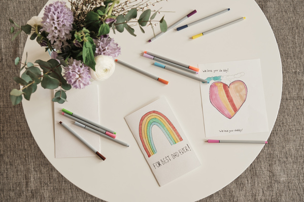 Handmade postcards for Fathers Day decorated with colorful images and applications and multi-colored markers are laid out on a white coffee table with a bouquet of flowers