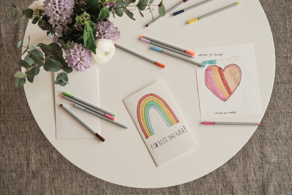 Flatlay of homemade Fathers Day cards decorated with bright images and applications with felt-tip pens is on a white coffee table with a bouquet of flowers
