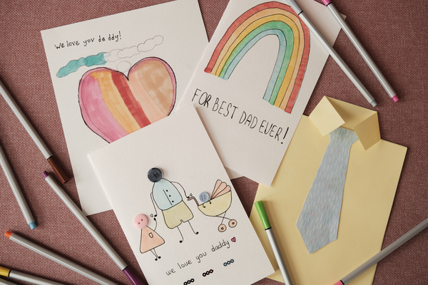 Handmade postcards dedicated to the Fathers holiday with bright illustrations and applications and multi-colored markers are scattered on the pink surface