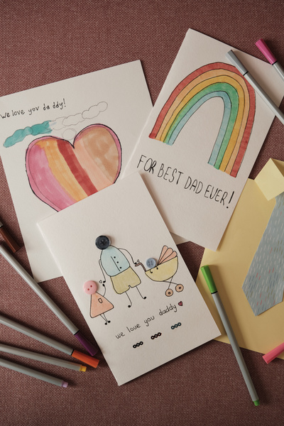 Handmade cards dedicated to the Fathers holiday decorated with colorful images and applications and multi-coloredfelt-tip pens are laid out on a pink surface