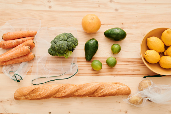 Top view of grocery purchases laid out on a wooden table such as vegetables with reusable bags citrus fruits in a bowl and a French baguette