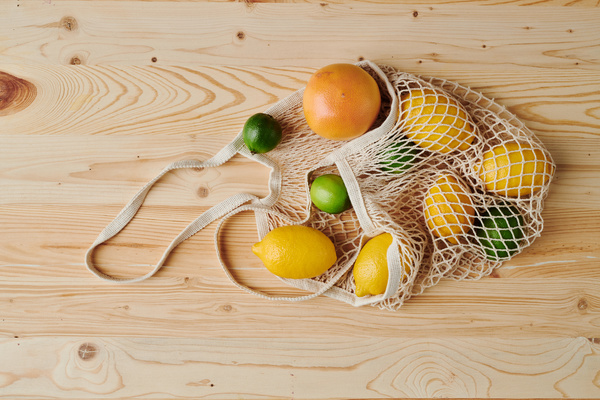 A white cotton string bag lying on a light wood table is filled with citrus fruits such as lime lemon and grapefruit